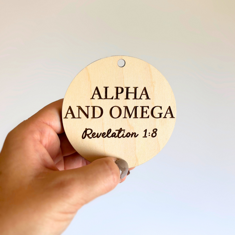 Hand holding a wooden ornament with 'Alpha and Omega, Revelation 1:8' text against a light background