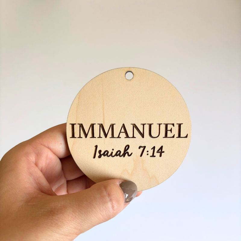 Round wooden ornament with 'IMMANUEL Isaiah 7:14' text held by a hand against a light background