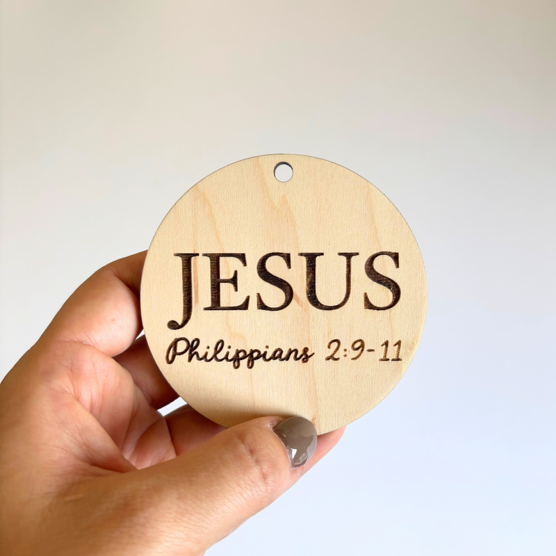 Hand holding a wooden ornament with 'JESUS' and 'Philippians 2:9-11' text.