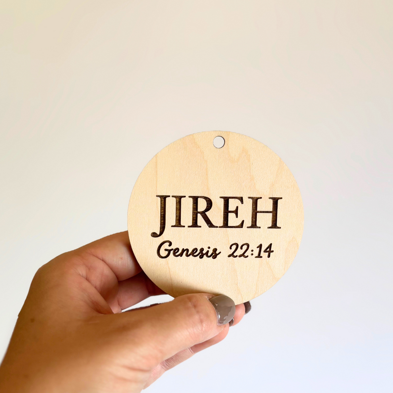 Hand holding a wooden ornament with 'JIREH' and 'Genesis 22:14' text against a plain background