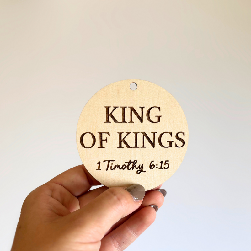 Round wooden sign with 'King of Kings 1 Timothy 6:15' text held by a hand against a light background