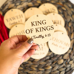 Hand holding a wooden round ornament with 'King of Kings' text, surrounded by similar ornaments.