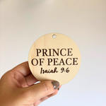 Round wooden sign with 'Prince of Peace' and Isaiah 9:6 text held by a hand against a light background