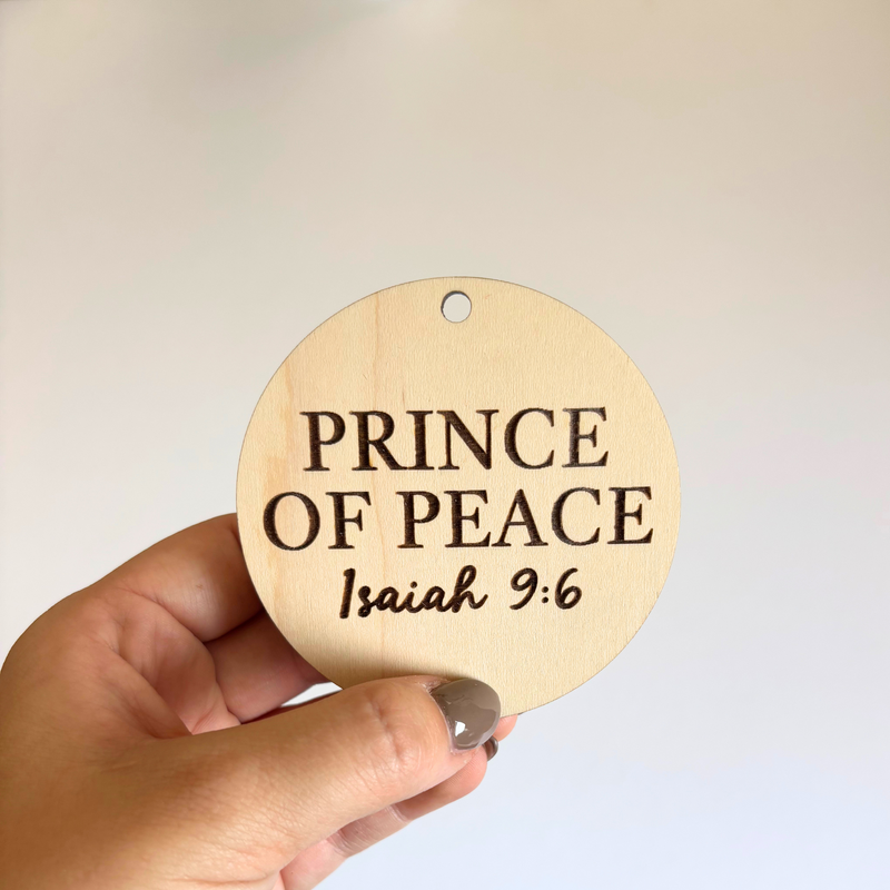 Round wooden sign with 'Prince of Peace' and Isaiah 9:6 text held by a hand against a light background