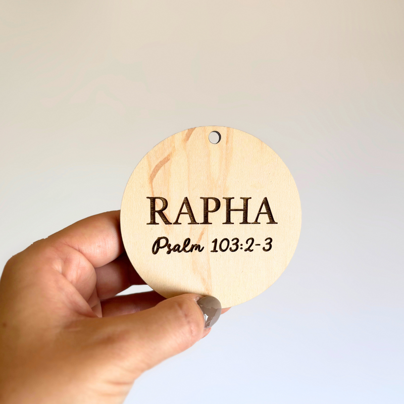 Hand holding a round wooden ornament with 'RAPHA Psalm 103:2-3' text on a plain background