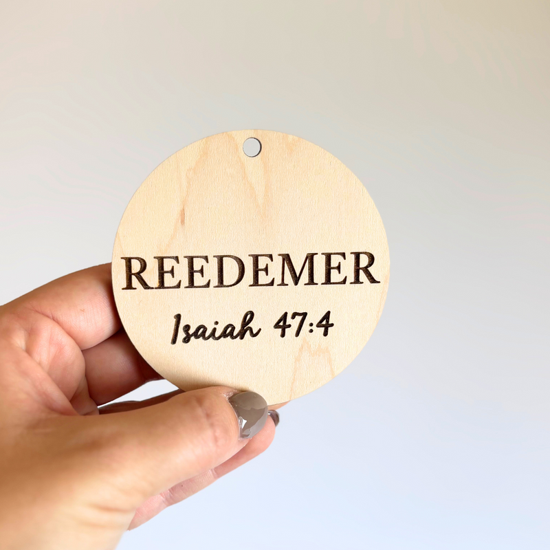 Hand holding a wooden round tag with 'REDEMER' and 'Isaiah 47:4' text against a light gray background