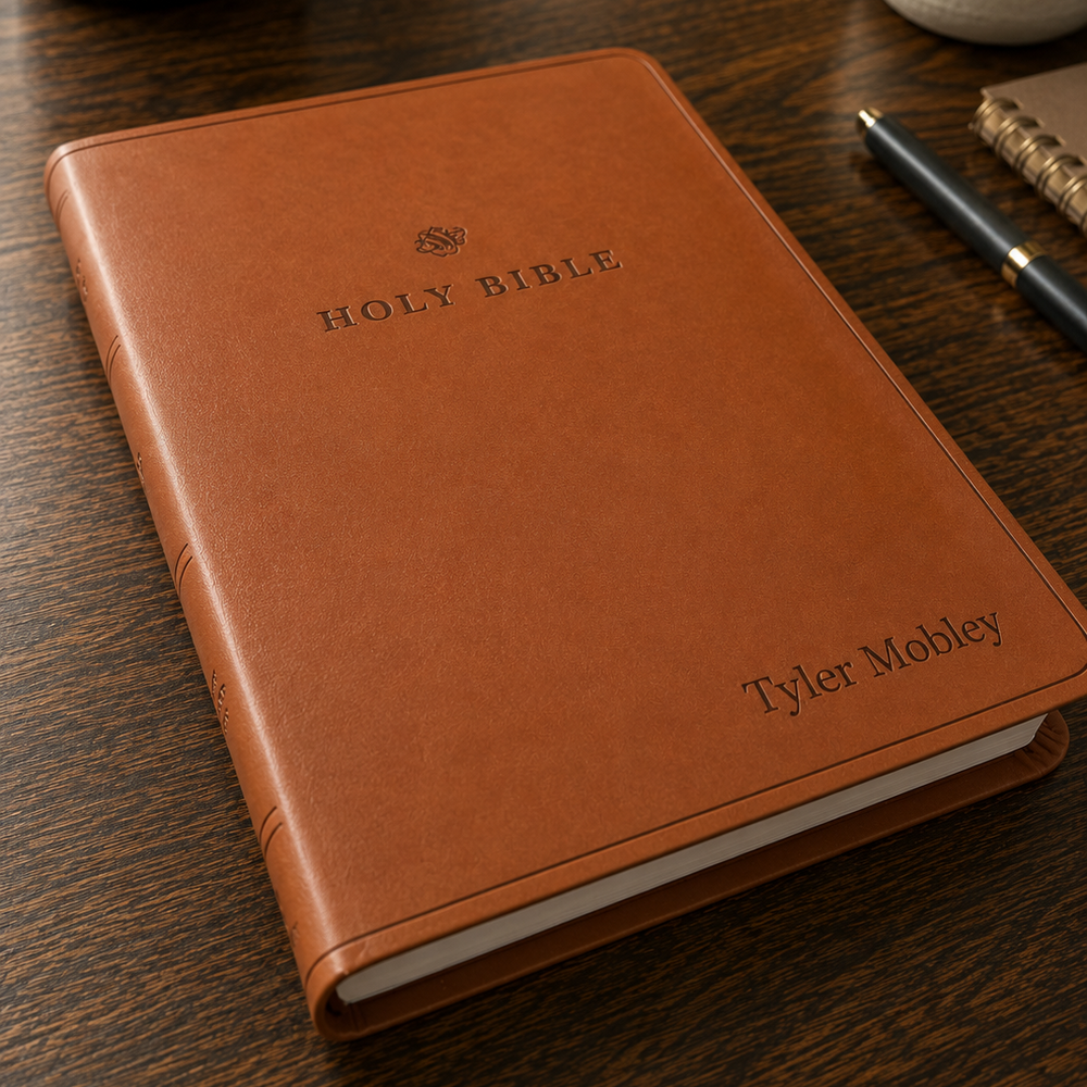 Brown leather Bible with 'Tyler Mobley' engraving on a wooden surface with a cup and plant in the background.