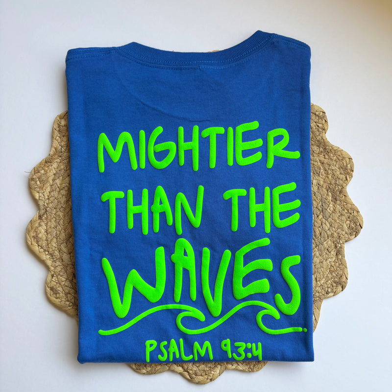 Blue t-shirt with green text 'Mightier Than The Waves Psalm 93:4' on a textured surface.